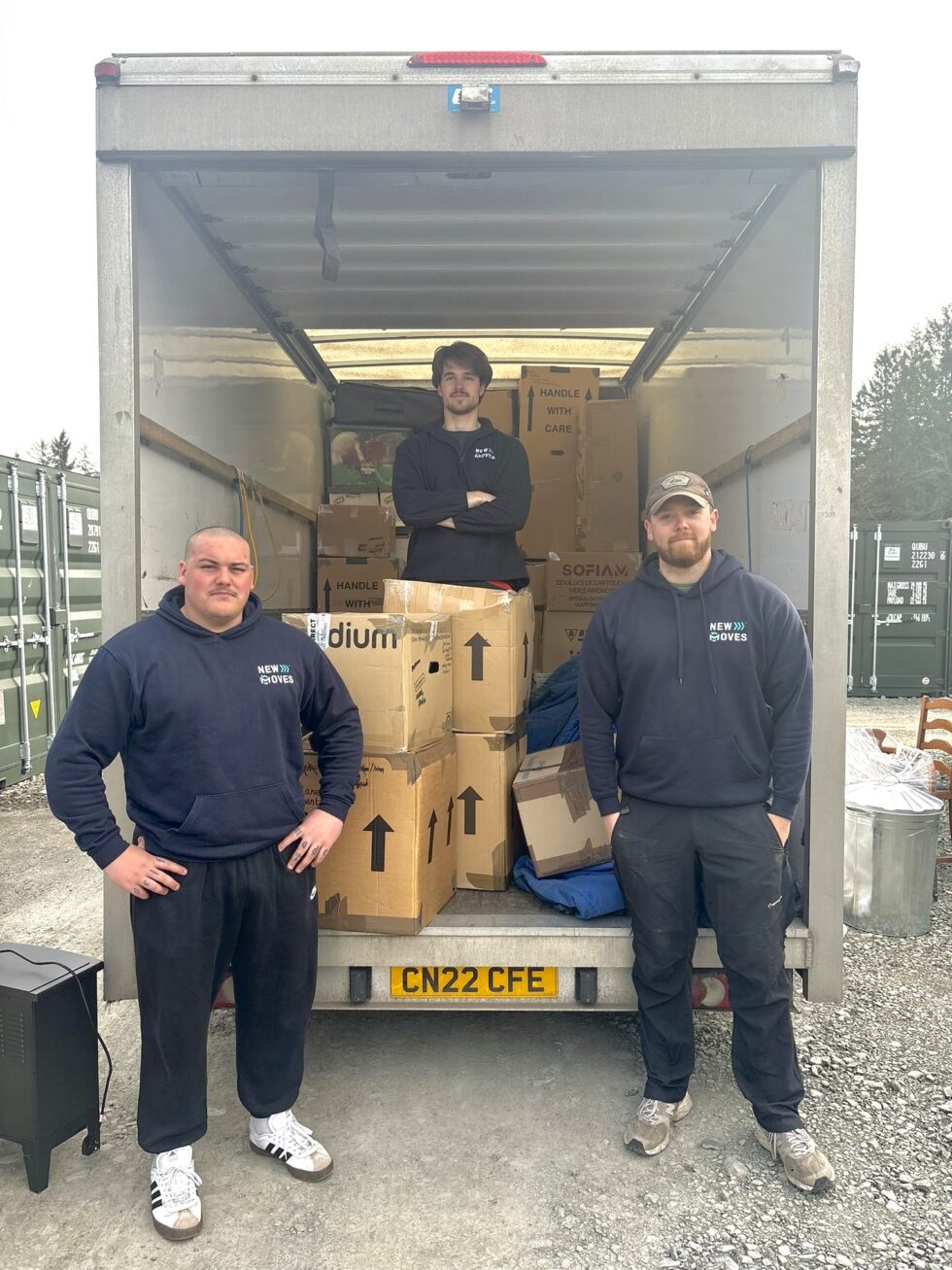 Three New Moves team members standing in front of an open removal van loaded with boxes and furniture at a storage facility