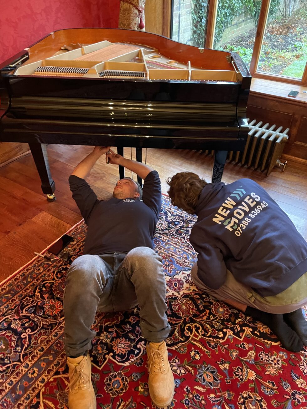 Two New Moves team members working underneath a black grand piano to prepare its legs for a move, on a Persian rug in a period room