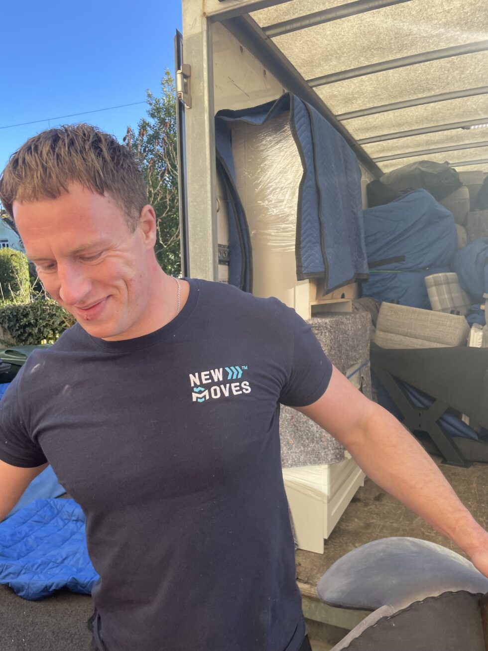 New Moves team member in a branded t-shirt in front of an open removal van packed with wrapped furniture and boxes